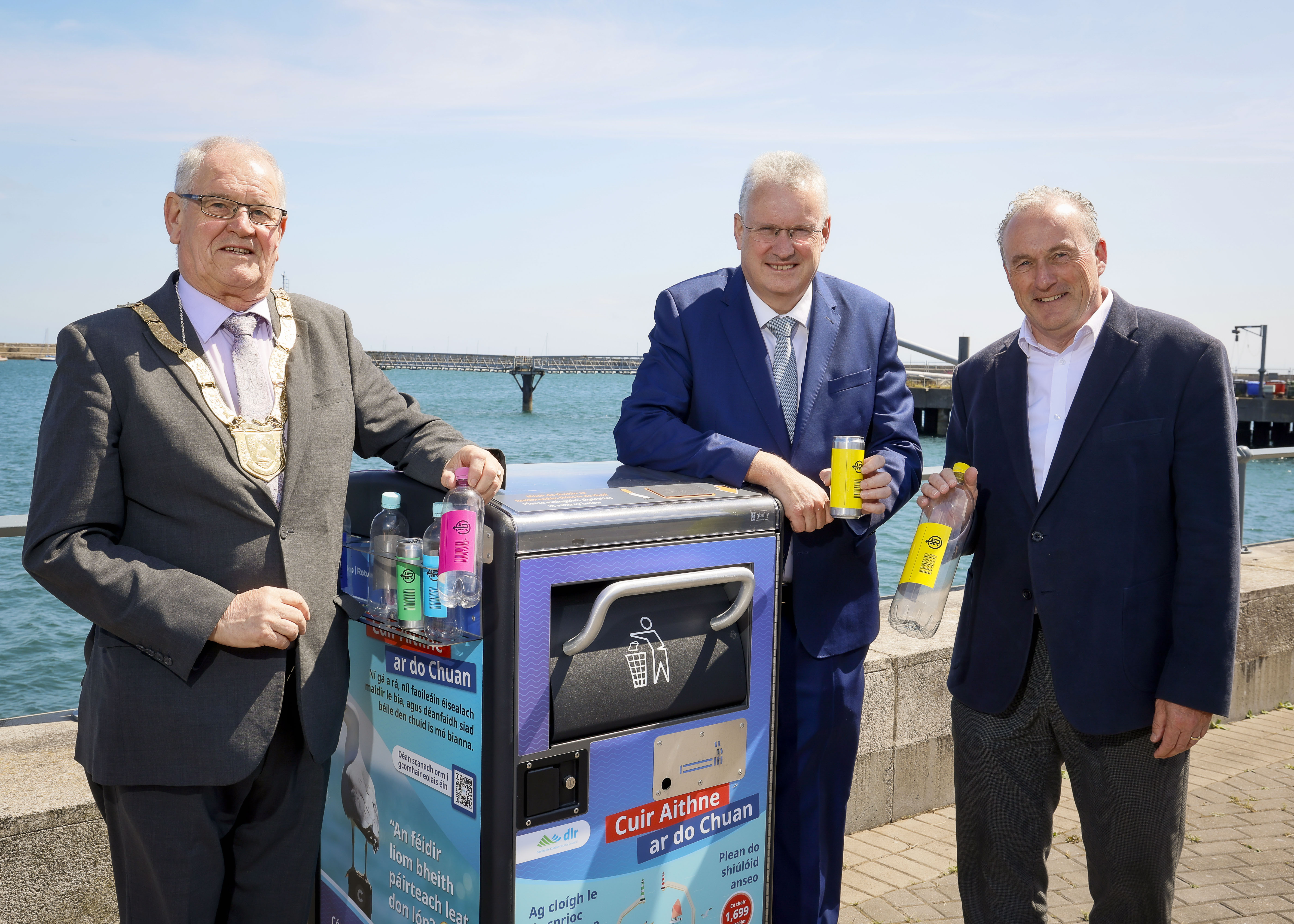 Three men standing beside a seaside recycling bin wrapped in Re-turn Deposit Return Scheme branding, holding plastic bottles and cans. The bin displays educational messaging about recycling and marine protection.
