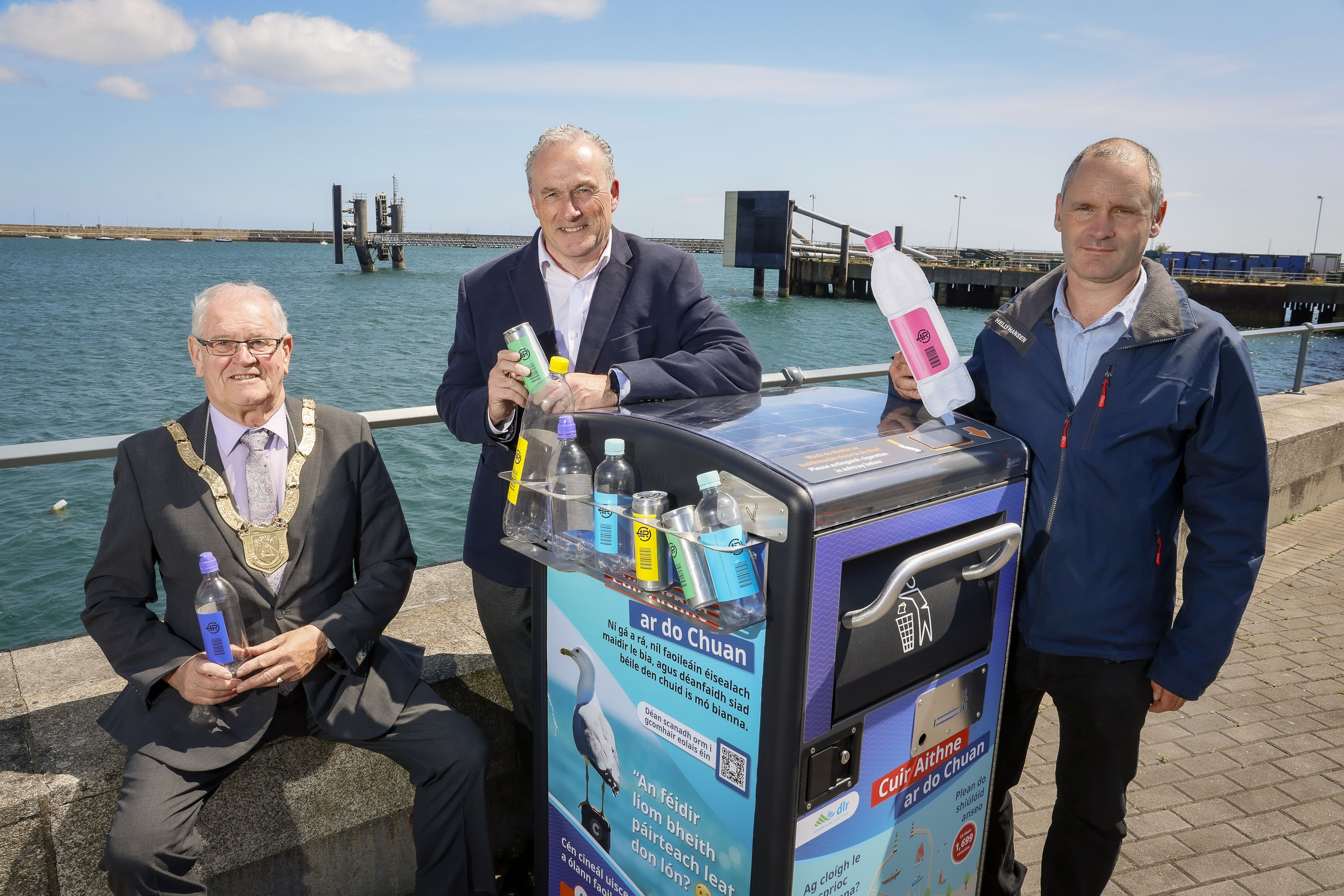 Three men standing beside a seaside recycling bin wrapped in Re-turn Deposit Return Scheme branding, holding plastic bottles and cans.