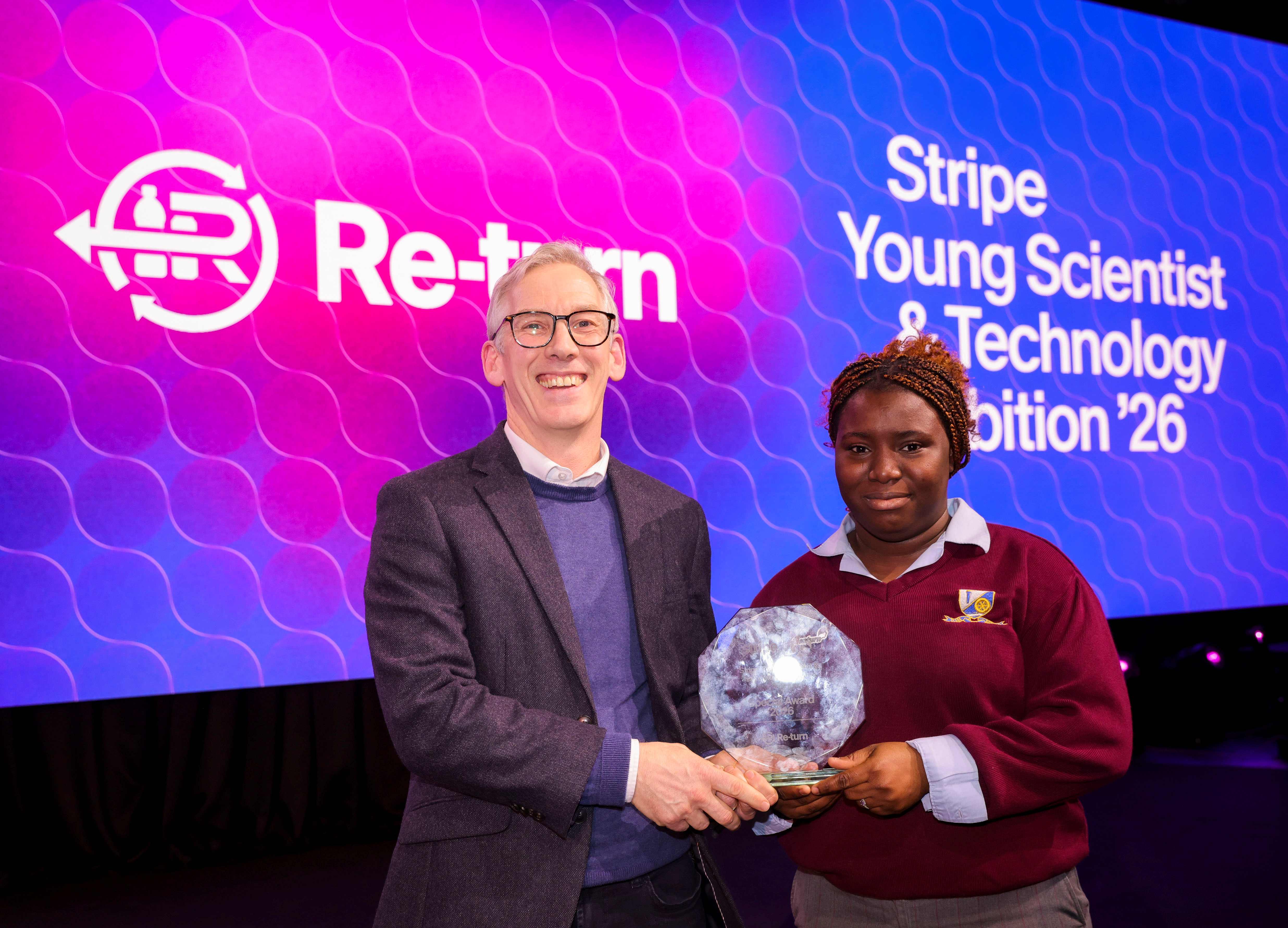 Two individuals standing on a stage holding a crystal-like award in front of a large screen displaying the Re-turn logo and the text ‘Stripe Young Scientist & Technology Exhibition ’26’.