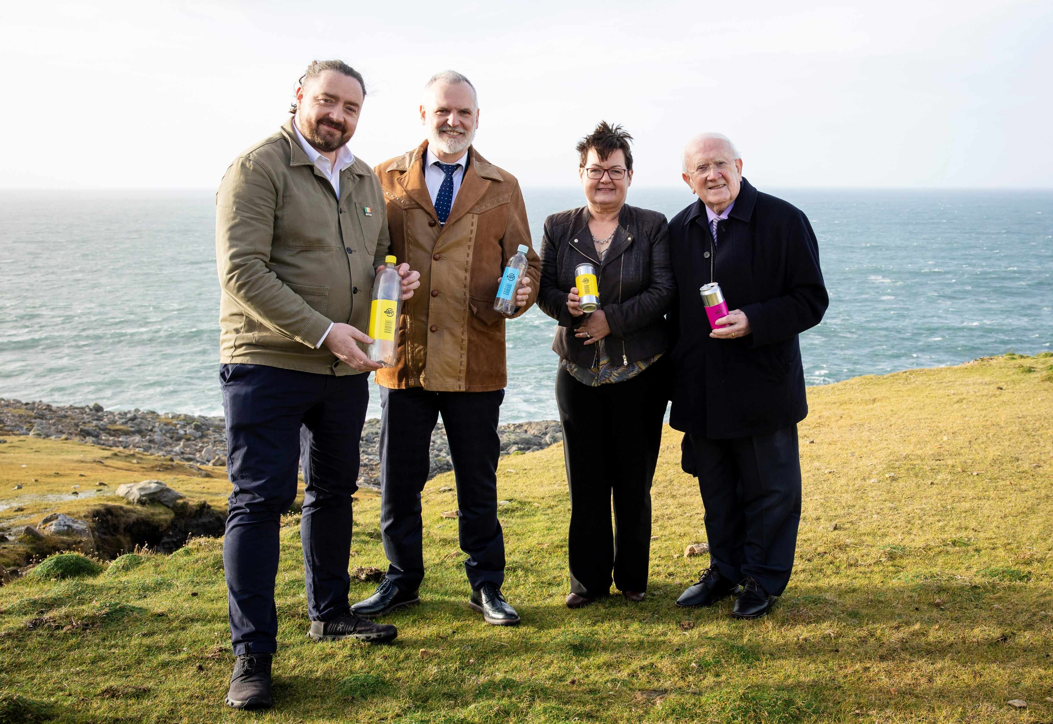 Four individuals standing outdoors on a grassy area near a rocky coastline, each holding colorful beverage containers. The ocean and horizon are visible in the background under a bright sky.