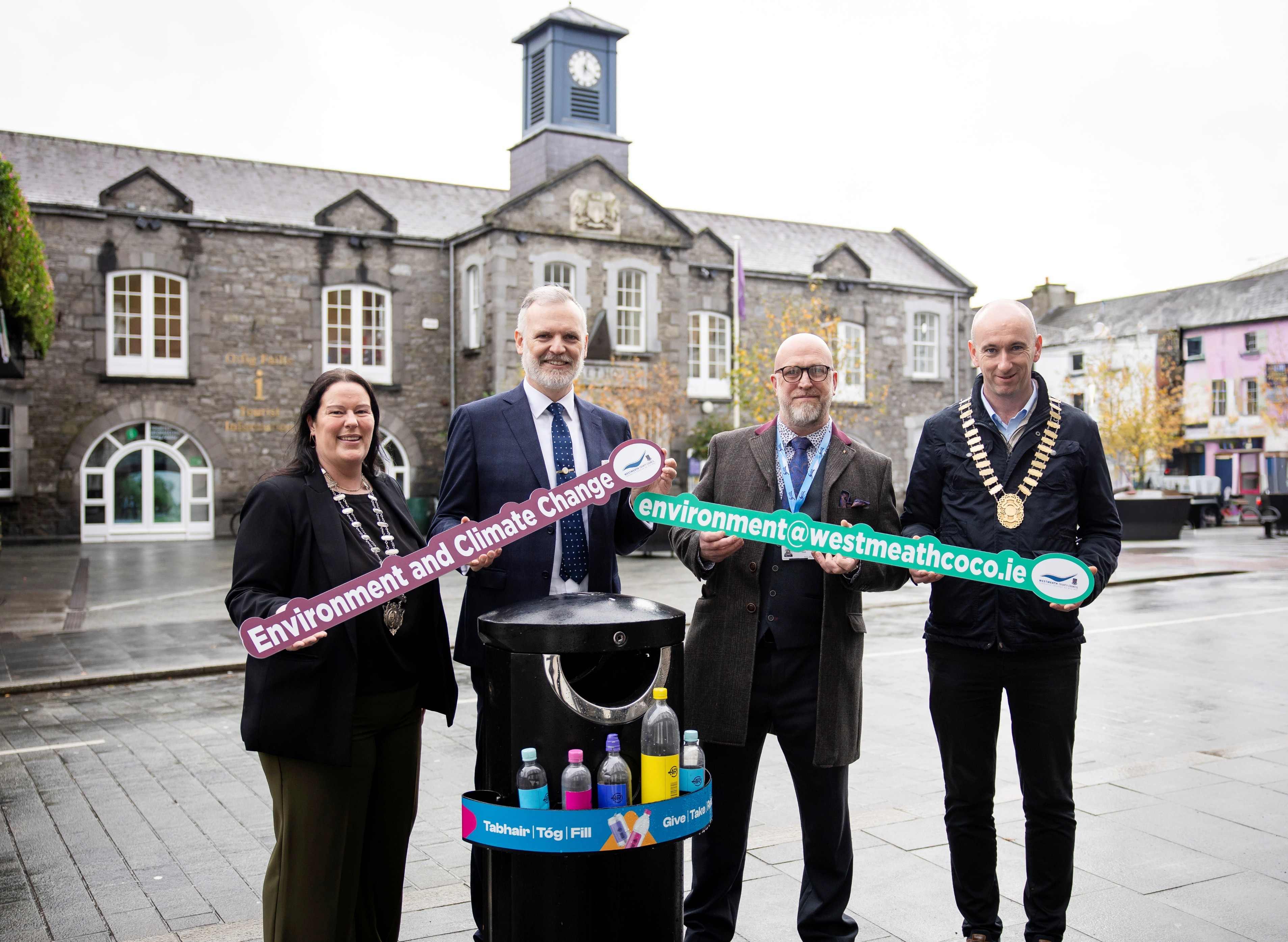 Four individuals standing outdoors in front of a stone building with a clock tower, holding two large banners that read ‘Environment and Climate Change’ and ‘environment@westmeathcoco.ie’. In front of them is a black recycling bin with several colorful plastic bottles displayed on top, featuring a label with text in Irish and English promoting recycling.