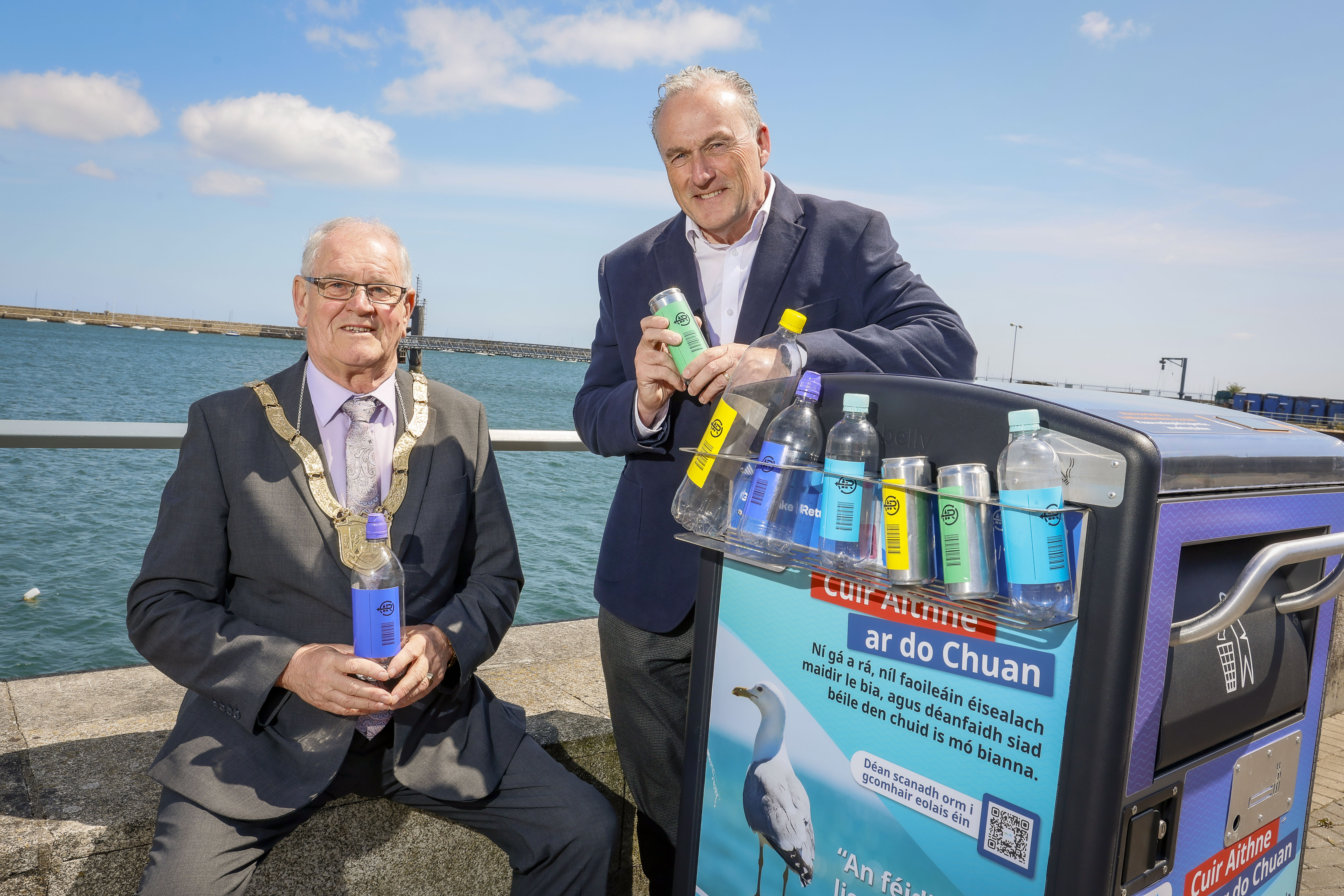 Cllr Jim Gildea, Cathaoirleach, Dún Laoghaire-Rathdown County Council & Ciaran Foley, CEO of Re-turn standing beside a seaside recycling bin wrapped in Re-turn Deposit Return Scheme branding, holding plastic bottles and cans.