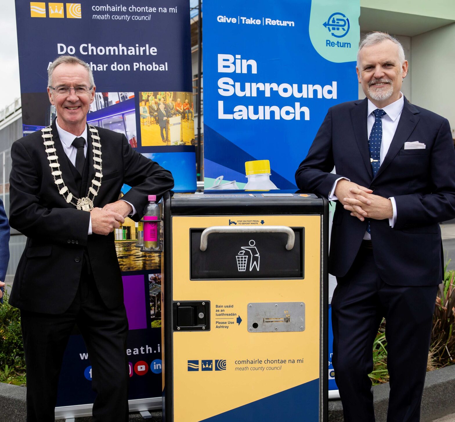 Three individuals standing in front of a bin with promotional banners for the 'Bin Surround Launch' initiative. The signage includes English and Irish text referencing Meath County Council and waste disposal. The setting suggests an official event promoting improved public bin infrastructure.