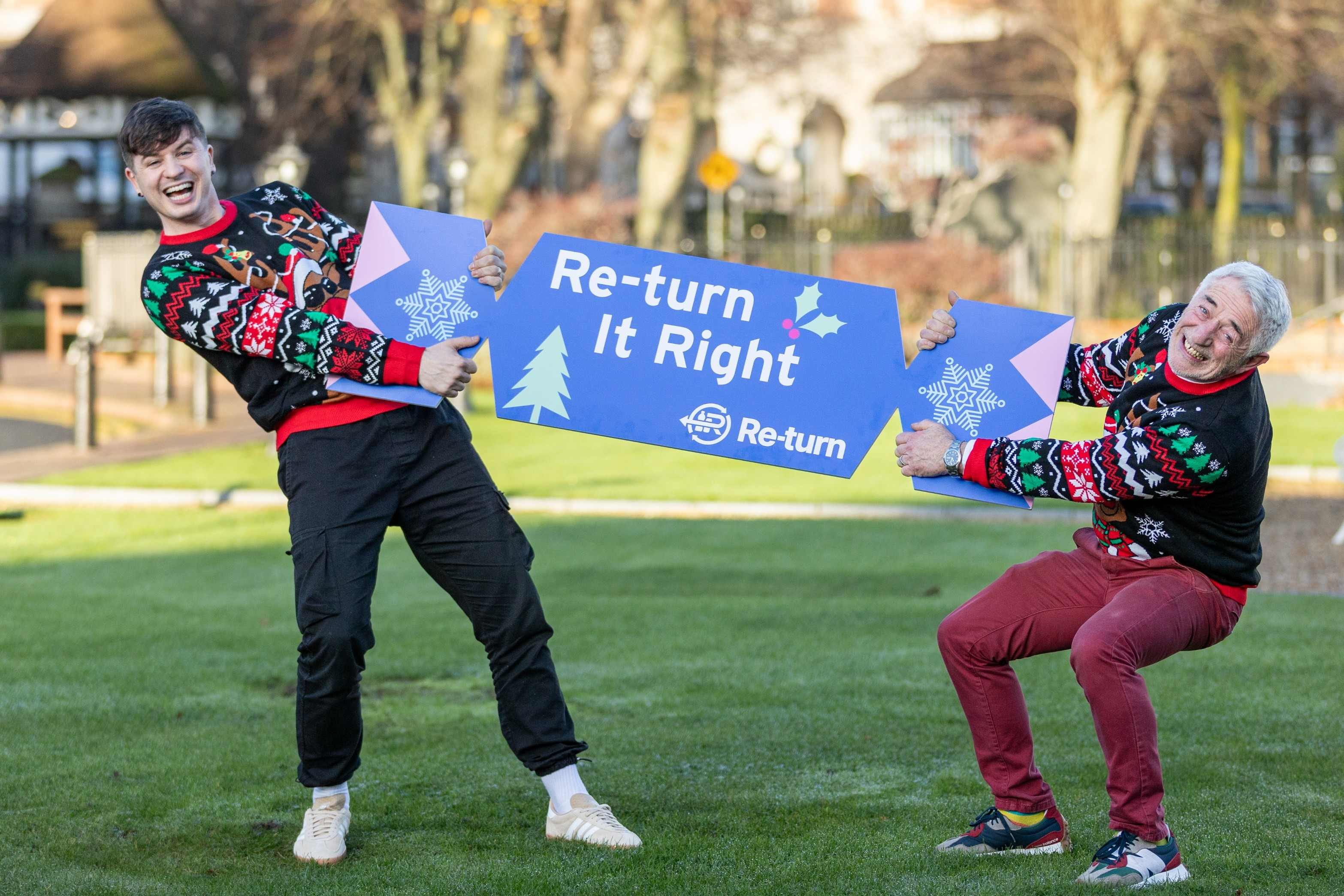 Two people wearing festive Christmas jumpers hold a large blue sign that reads ‘Re-turn It Right’ with snowflake and tree graphics, standing on a grassy area outdoors.